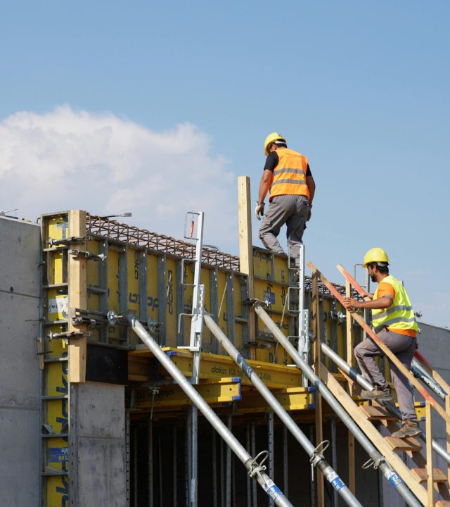 Two construction workers building a structure, embodying teamwork and safety under a clear blue sky.